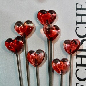May include: Close-up of several heart-shaped glass ornaments on metal stems. The hearts are a translucent red with darker red and white accents. The stems are thin and gray, and the background is a light gray.