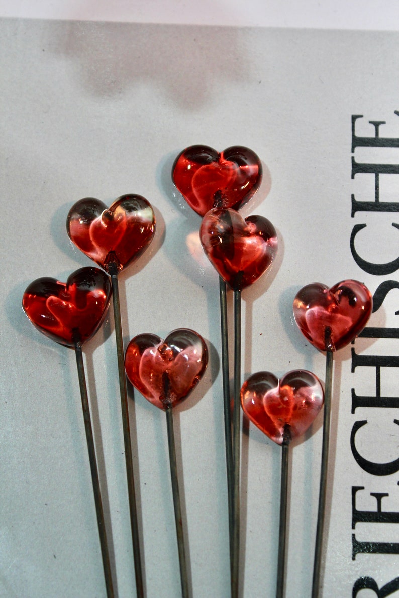 May include: Close-up of several heart-shaped glass ornaments on metal stems. The hearts are a translucent red with darker red and white accents. The stems are thin and gray, and the background is a light gray.