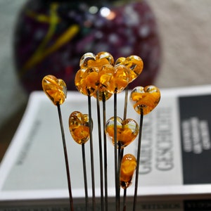 May include: A collection of heart-shaped glass ornaments on slender black stems. The transparent hearts are filled with a golden-yellow material. The ornaments are arranged in front of a book and a blurred purple vase.
