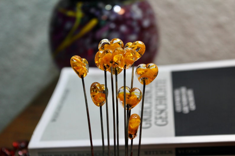 May include: A collection of heart-shaped glass ornaments on slender black stems. The transparent hearts are filled with a golden-yellow material. The ornaments are arranged in front of a book and a blurred purple vase.