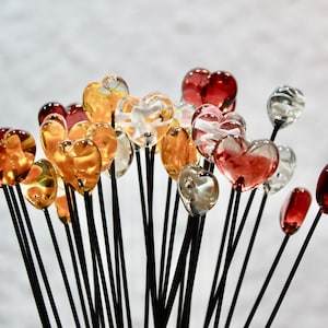 May include: A collection of glass heart-shaped ornaments in various colors, including red, amber, and clear. Each heart is mounted on a black stem, creating a bouquet-like arrangement. The hearts vary in size and transparency.