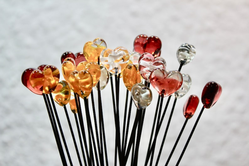 May include: A collection of glass heart-shaped ornaments in various colors, including red, amber, and clear. Each heart is mounted on a black stem, creating a bouquet-like arrangement. The hearts vary in size and transparency.
