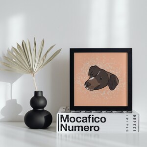 Puede incluir: Una impresión enmarcada en negro de la cabeza de un perro marrón y blanco sobre un fondo melocotón con motas blancas. La impresión está encima de un libro blanco con el texto "Mocacifico Numero" en negro.