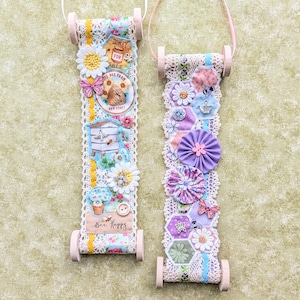 May include: Two decorative hanging ornaments with floral and bee-themed embellishments. Each ornament features a fabric roll design with lace, flowers, and a handle. One has a "Bee Happy" sign. The color palette includes pink, white, yellow, and blue.