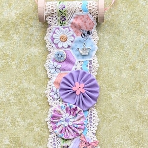 May include: A decorative hanging ornament featuring a series of hexagonal fabric patches in various colors, including purple, pink, and blue. The patches are adorned with floral and butterfly embellishments, and are connected by a lace trim. The ornament is suspended by a pink ribbon.