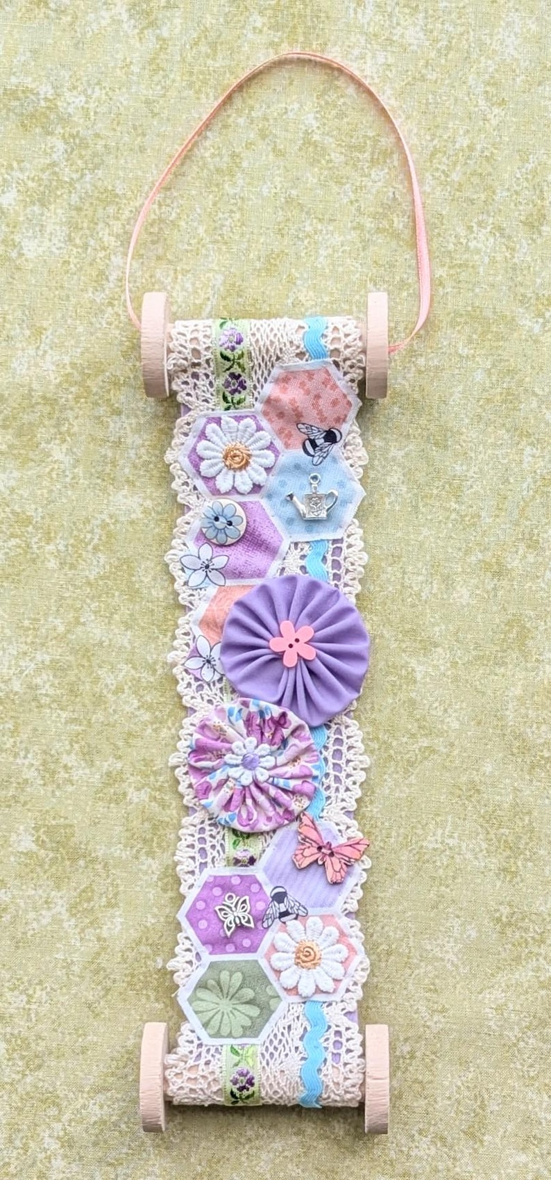 May include: A decorative hanging ornament featuring a series of hexagonal fabric patches in various colors, including purple, pink, and blue. The patches are adorned with floral and butterfly embellishments, and are connected by a lace trim. The ornament is suspended by a pink ribbon.