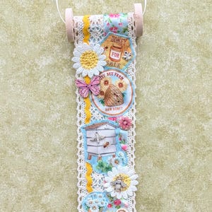 May include: A decorative hanging scroll featuring a bee theme. It includes floral and bee-related imagery, such as a beehive, flowers, and the text "Honey for Sale" and "Bee Happy". The scroll is trimmed with lace and has wooden dowels at the top and bottom.