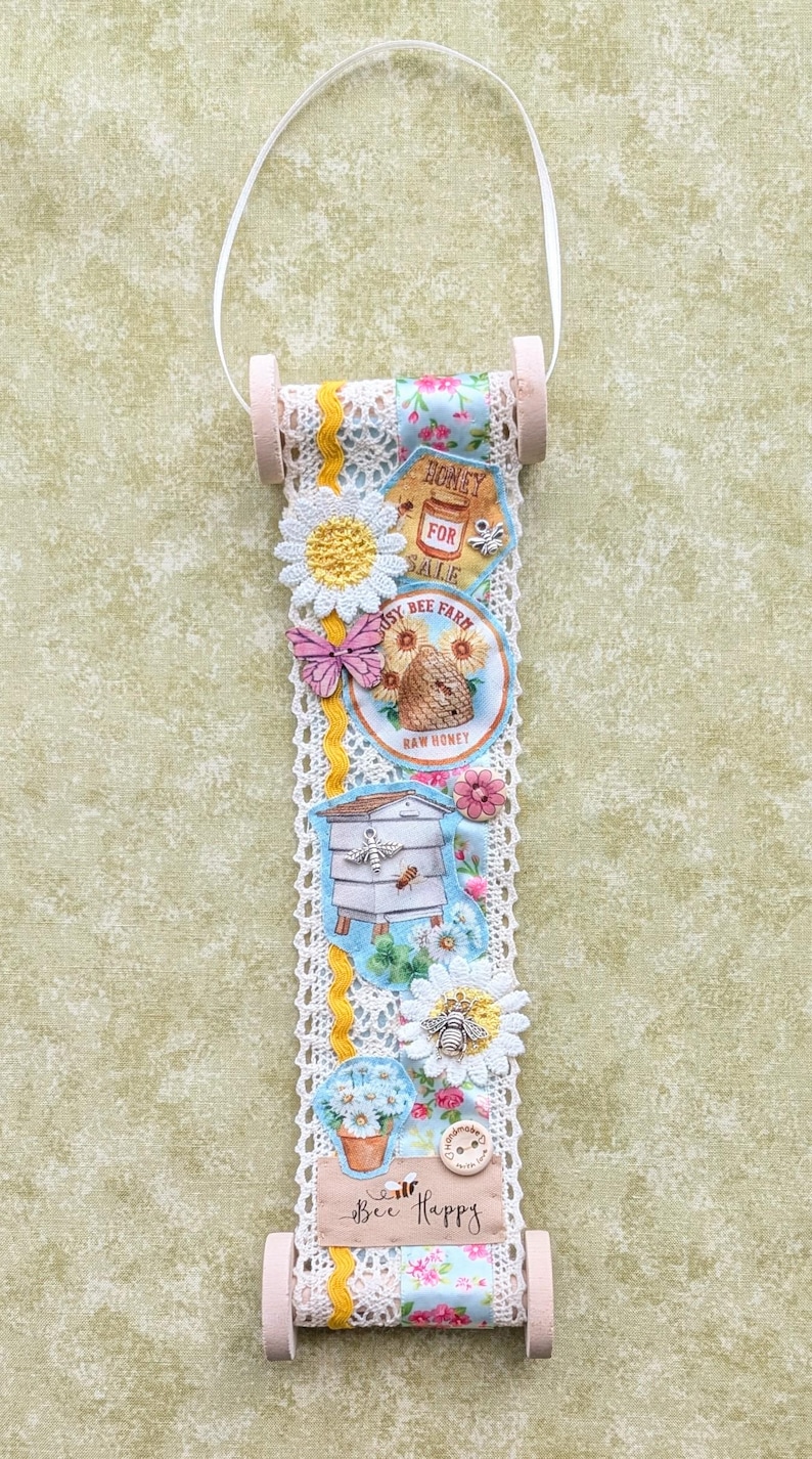 May include: A decorative hanging scroll featuring a bee theme. It includes floral and bee-related imagery, such as a beehive, flowers, and the text "Honey for Sale" and "Bee Happy". The scroll is trimmed with lace and has wooden dowels at the top and bottom.