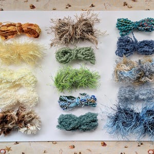 May include: A collection of fifteen assorted yarn bundles arranged in three columns on a white surface. The yarns feature various textures including fuzzy, metallic, ribbon-like, and twisted strands in shades of brown, yellow, green, and blue.