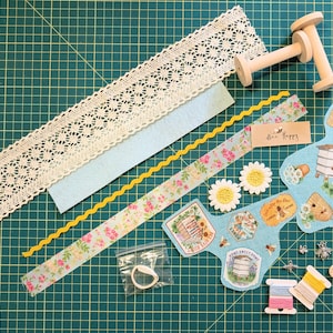 May include: A collection of craft supplies on a green grid surface. Includes lace trim, light blue felt, floral ribbon, yellow rickrack, wooden spools, fabric patches with bee and hive designs, daisy embellishments, buttons, thread spools, and small charms.