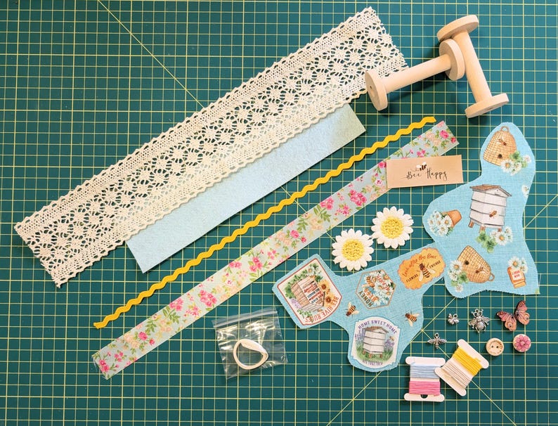 May include: A collection of craft supplies on a green grid surface. Includes lace trim, light blue felt, floral ribbon, yellow rickrack, wooden spools, fabric patches with bee and hive designs, daisy embellishments, buttons, thread spools, and small charms.
