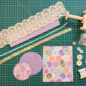 May include: A collection of craft supplies including lace trim, felt, ribbon, paper, wooden spools, buttons, and fabric. The items are arranged on a green cutting mat, suggesting a crafting or DIY theme. Various colors and textures are visible.