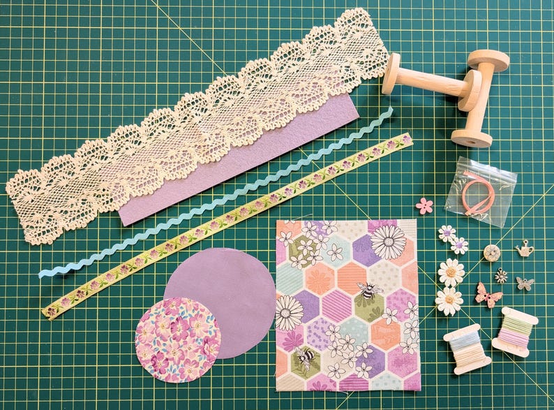 May include: A collection of craft supplies including lace trim, felt, ribbon, paper, wooden spools, buttons, and fabric. The items are arranged on a green cutting mat, suggesting a crafting or DIY theme. Various colors and textures are visible.