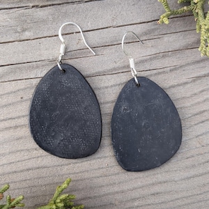 May include: A pair of black oval-shaped earrings with silver hooks. The earrings are made of a textured material.