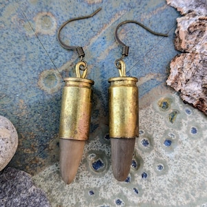 May include: A pair of gold bullet casing earrings with a brown wood accent at the bottom. The earrings have a simple hook closure.