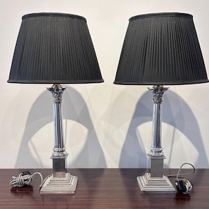 Peut inclure: Deux lampes de table assorties avec des abat-jours plissés noirs. Les lampes ont des bases argentées en forme de colonne avec des détails décoratifs. Elles sont posées sur une surface en bois foncé.