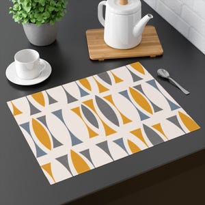 Peut inclure: Un set de table rectangulaire avec un motif répété de formes abstraites jaunes, grises et bleues sur fond blanc cassé. Le set de table est posé sur une surface sombre, avec une tasse et une soucoupe, une théière et une cuillère.