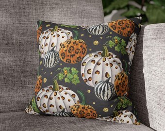 May include: Square decorative pillow with a dark gray background featuring a pattern of white and orange pumpkins with leopard print spots, green leaves, and gold glitter accents. The pillow is placed on a gray sofa.