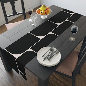 Op de afbeelding: Een zwart-wit geometrische tafelloper op een donkere houten tafel. De tafel is gedekt met een wit bord, bestek, wijnglazen en een kom met appels. De loper voegt een moderne touch toe aan de eethoek.