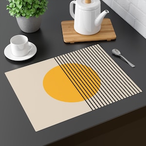May include: A beige placemat with a geometric design featuring a yellow sun shape and black vertical lines. A white teacup and saucer, a teapot, and a spoon are on a dark surface.