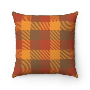 May include: Square throw pillow with a plaid pattern in shades of orange, red, and brown. The pillow has a repeating square design with diagonal lines within each square. The pillow is a decorative item for a home.