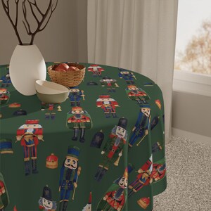 Peut inclure: Une nappe ronde vert foncé avec un motif répété de casse-noisettes colorés. La table est ornée d'un vase blanc, d'un petit bol et d'un panier en osier rempli de pommes rouges. La nappe mesure environ 152 cm de diamètre.