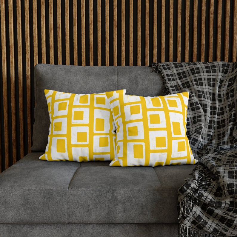 Pu&ograve; includere: Due cuscini con un motivo geometrico di quadrati gialli su sfondo bianco. I cuscini sono su un divano grigio con una coperta a quadri in bianco e nero.