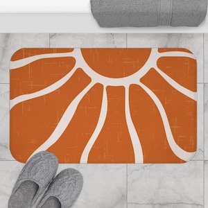 Puede incluir: Una alfombra de baño rectangular con un diseño retro. La alfombra presenta un fondo naranja quemado con un patrón de sol blanco. Dos zapatillas de punto gris están en la parte inferior de la alfombra. Una toalla gris está en la parte superior.