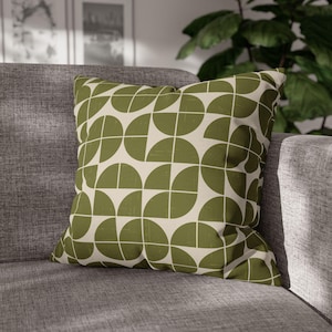 May include: Square decorative pillow with a geometric pattern of olive green semi-circles on a cream background. The pillow is placed on a gray sofa, offering a modern touch to home decor.
