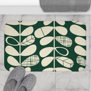 May include: A rectangular bath mat with a dark green background and a cream-colored leaf pattern. The leaves have various geometric designs. Gray slippers are at the bottom left. A gray towel is at the top.