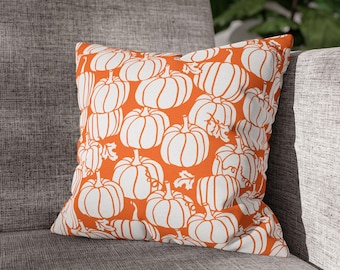 May include: A square cushion featuring a repeating pattern of white pumpkins on an orange background. The cushion is placed on a grey sofa, suggesting an autumn theme. The design is simple and modern, suitable for home decor.