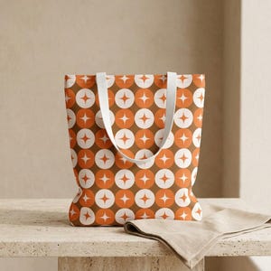 Puede incluir: Un bolso tote con un patrón retro de círculos naranjas, blancos y marrones con estrellas. El bolso tiene asas blancas y está sobre una superficie clara. Una servilleta beige está al lado del bolso.
