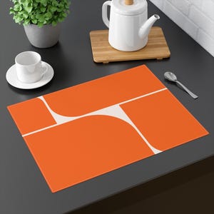 May include: An orange placemat with a white geometric design. The placemat features rectangular and curved shapes in a modern, minimalist style. The placemat is on a dark surface, with a white teacup and saucer, a teapot, and a spoon.