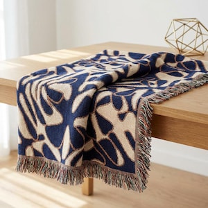 May include: A navy and beige patterned throw blanket with a fringed edge is displayed on a wooden table. The blanket's design is abstract. A gold geometric ornament is also on the table.