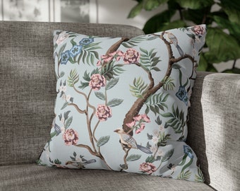 May include: Square cushion with a light blue background featuring a floral and bird design. The pattern includes pink flowers, green leaves, and brown branches. The cushion is placed on a grey sofa.