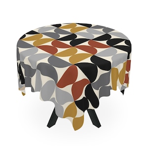 May include: A round tablecloth featuring an abstract pattern in grey, black, rust, and gold on a cream backdrop. The cloth is draped over a black table with three legs. The table is likely to be around 100 cm in diameter.