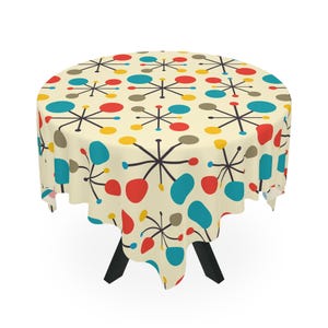 May include: A round tablecloth with a mid-century modern design. The cream-coloured fabric features a repeating pattern of black starbursts, red, blue, yellow, and grey shapes. The tablecloth is draped over a black table.