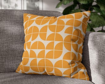 May include: Square cushion featuring a geometric design of orange semi-circles and white lines on a cream backdrop. The cushion is placed on a grey sofa, offering a modern aesthetic. This is a home decor item, perfect for adding a touch of style.