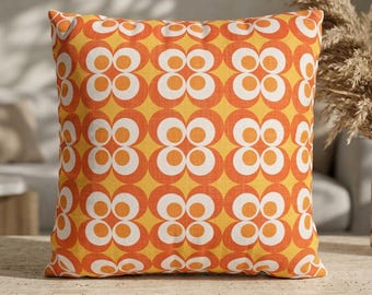 May include: A square throw pillow featuring a repeating geometric pattern of stylised four-petal flowers. The design consists of vibrant orange and white circular shapes set against a warm yellow background, creating a bold, symmetrical visual effect.