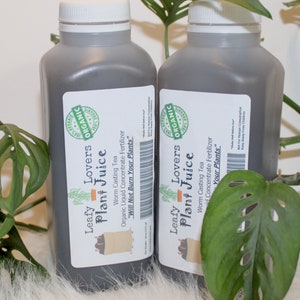 May include: Two dark gray plastic bottles with white lids and labels. The labels are white with black text and a green illustration of a plant. The text on the labels reads "Leafy Lovers Plant Juice" and "Worm Casting Tea Organic Liquid Concentrate Fertilizer "Will Not Burn Your Plants".