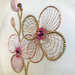 May include: A delicate floral embroidery design featuring pink and gold flowers with sparkling pink and purple rhinestones. The embroidery is on a white fabric background.