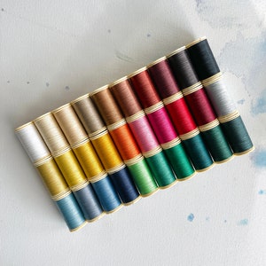 May include: A collection of 20 spools of thread in various colors, including white, beige, yellow, orange, red, pink, green, blue, and black. The spools are arranged in a row, with the colors transitioning from light to dark.