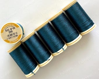 Op de afbeelding: Een bovenaanzicht toont zes spoelen teal zijden draad. Eén spoel staat apart, de andere vijf staan op een rij. De spoelen hebben een lichtbeige basis en zijn voorzien van Franse tekst.