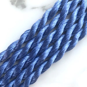 May include: Close-up of several strands of twisted, periwinkle-colored embroidery floss. The floss has a slight sheen, and the strands are tightly wound together. The background is a white, marble-like surface.