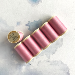 May include: Six spools of pink thread arranged diagonally on a white surface with blue watercolor accents. One spool is separate, while the others are grouped together. The spools have a light beige base and are labeled with text.