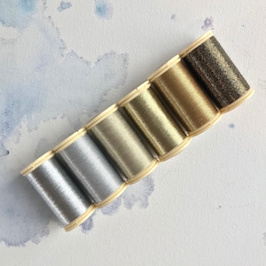 May include: Six spools of metallic thread in various shades of gold and silver. The spools are arranged in a row from darkest to lightest.