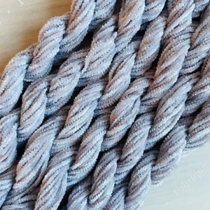 May include: Close-up of a braided, light gray, plush yarn. The yarn is thick and soft, with a velvety texture. The braid is tightly woven, creating a textured surface. The overall effect is cozy and inviting, suitable for crafting projects.