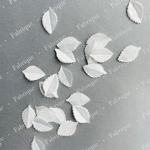 May include: A collection of white fabric leaves scattered on a gray surface. The leaves are delicate and have a slightly textured appearance.