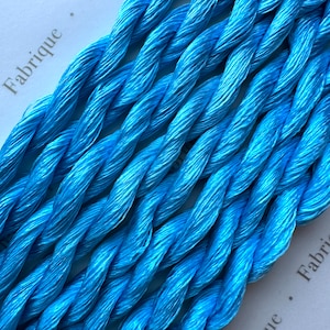 May include: Close-up of a bundle of light blue embroidery floss. The floss is twisted and has a slight sheen. The text "Fabrique" is visible on the white background.