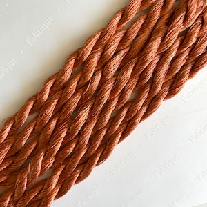 May include: Close-up of a bundle of six strands of embroidery floss in a warm, reddish-brown color. The floss is arranged in a diagonal pattern, with the ends of the strands visible at the top and bottom of the image. The floss is made of a soft, silky material and has a subtle sheen.